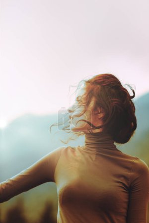 young woman gazes to the side, her hair tousled by the wind. She wears a brown turtleneck, and the background features soft, blurred colors and distant hills under a pastel sky