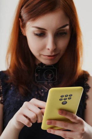 Young woman with auburn hair intently looks at her yellow smartphone, which has an unusually high number of cameras on the back. She wears a dark lace top and is focused on the screen
