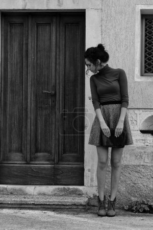 Eine junge Frau steht vor einer Holztür und schaut zur Seite. Sie trägt einen braunen Rollkragen und einen grauen Rock. Ihre Hände ruhen auf einer Clutch, und die Szene ist schwarz-weiß