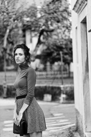 young woman stands near a building, looking at something away. She wears a brown turtleneck and a grey skirt, holding a clutch. The black and white photo features blurred trees in the background