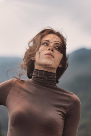 young woman tilts her head back, wind blowing her hair across her face. Her expression is calm and reflective. She wears a brown turtleneck, and the blurred background features distant hills