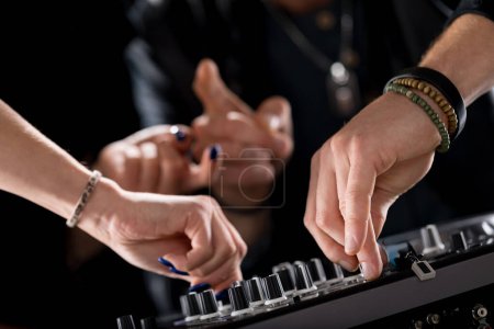 close-up of hands working on a DJ mixer, adjusting knobs, while other hands make gestures in the background. The scene emphasizes precision, control, and teamwork in a dynamic DJ environment