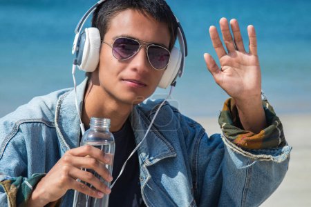young man with headphones drinking water relaxed on the beach and waving