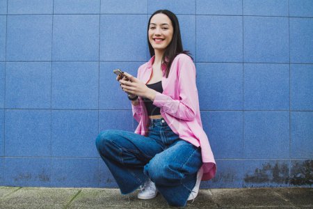 Foto de Chica urbana en cuclillas con teléfono móvil en la pared - Imagen libre de derechos