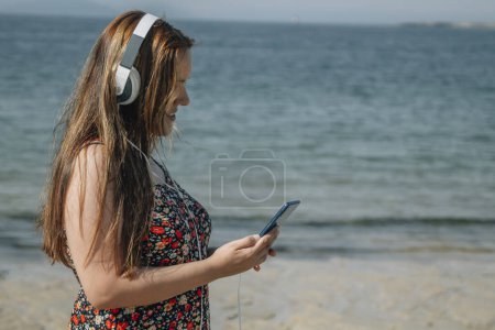 Téléchargez les photos : Belle femme plus la taille sur la plage avec téléphone et écouteurs - en image libre de droit