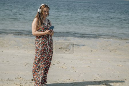 Téléchargez les photos : Belle femme plus la taille sur la plage avec téléphone et écouteurs - en image libre de droit
