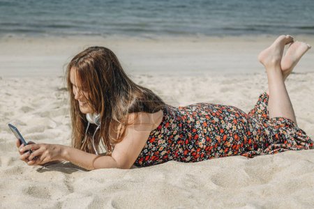 Téléchargez les photos : Belle femme plus la taille de détente sur la plage avec téléphone et écouteurs - en image libre de droit