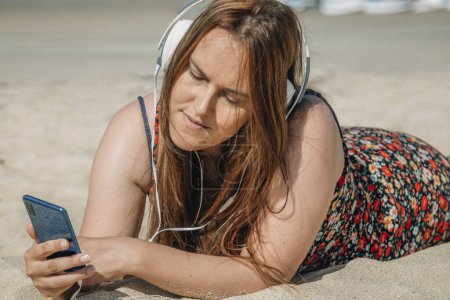 Téléchargez les photos : Femme courbée détendue sur la plage avec téléphone et écouteurs - en image libre de droit