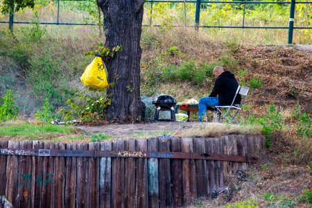 Téléchargez les photos : Berlin, Berlin / Allemagne - 09 03 2022 : Un homme qui a une chaise et une table sur les rives de la Spree, a accroché un sac poubelle sur un arbre et grille avec une bouilloire grill. - en image libre de droit