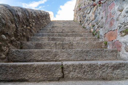 Foto de Una escalera de piedra que conduce hacia arriba, forrada por una pared de piedra alta a la derecha y una pared plana a la izquierda. En la parte superior se puede ver un cielo azul con nubes - Imagen libre de derechos