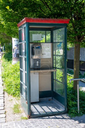 Foto de Salzburgo, Salzburgo - Austria - 17-06-2021: Cabina telefónica verde con techo rojo, de cristal y metal, sin puerta. En el interior, un teléfono y un estante son visibles. Sol brillante. - Imagen libre de derechos