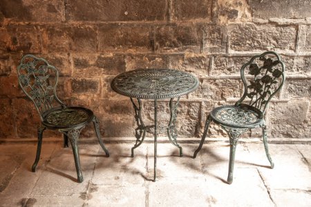 Téléchargez les photos : Une table et deux chaises en fer forgé avec des éléments finement fabriqués. Le mobilier semble très classique et se tient devant un mur de grandes vieilles pierres. - en image libre de droit
