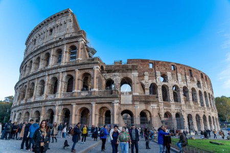 Foto de Roma, Lacio - Italia - 23-11-2022: La fachada exterior del Coliseo de Roma con muchos turistas en primer plano bajo un cielo azul. A la derecha se puede ver la hierba. - Imagen libre de derechos