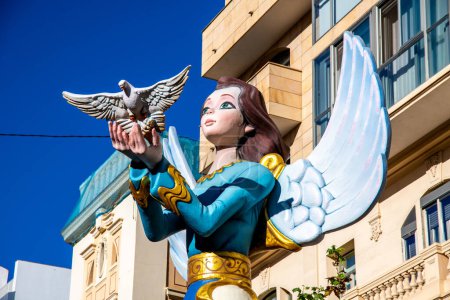 Foto de Alicante, Alicante - España - 01-07-2024: Vibrante efigie de Hogueras de San Juan, estatua de ángel agarrando una paloma, contra el cielo azul de Alicante, encarnando el espíritu del festival - Imagen libre de derechos
