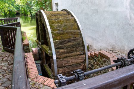 Foto de Wusterhausen, Brandenburgo - Alemania - 28-06-2022: Una vieja rueda de molino que ya no funciona en una pared de la casa y un río de madera y un mecanismo para regular el flujo de agua, llamado vertedero. - Imagen libre de derechos