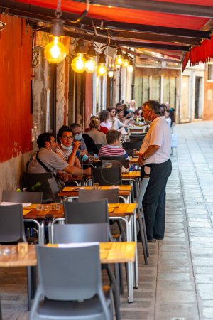 Foto de Venecia, Véneto - Italia - 06-10-2021: Los comensales disfrutan de comidas en un restaurante al aire libre a lo largo de una estrecha calle veneciana - Imagen libre de derechos