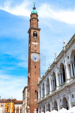 Vicenza, Venetien - Italien - 06.12.2021: Der ikonische rote Backstein Torre Bissara mit seiner einzigartigen Uhr und die weiße Basilika Palladiana bilden einen reizvollen Kontrast zum Himmel von Vicenza