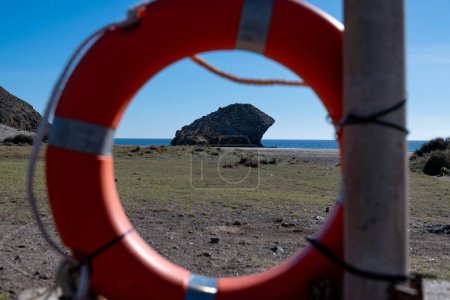 Cabo de Gata, Almeria - Spanien - 23.01.2024: Ein roter Rettungsring umrahmt eine entfernte Felsformation am Meer von Playa de Monsul in Cabo de Gata unter heller Sonne