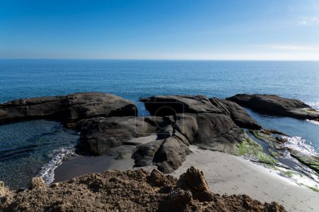 Cabo de Gata, Almería - España - 23-01-2024: Las suaves olas bañan las rocas cubiertas de algas en una soleada costa de Cabo de Gata