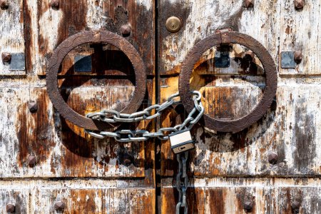 Photo for Vicenza, Venetien - Italy - 06-12-2021: A shiny chain and silver padlock secure a weathered wooden door with rusty iron rings, combining old and modern - Royalty Free Image