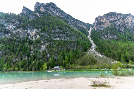 Alpen, Südtirol - Italien - 06-07-2021: Haus an einem türkisfarbenen See mit breitem, sandigem Ufer vor imposanten, hoch aufragenden Felsen mit grünem Kiefernwald in Südtirol