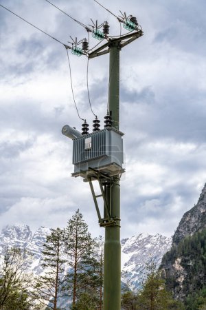 Alpen, South Tyrol - Italy - 06-07-2021: Power line tower stands amidst snowy mountain peaks and coniferous trees against a white sky backdrop