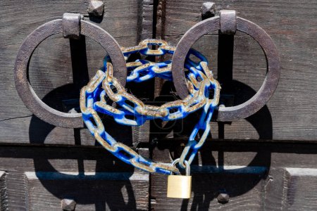 Photo for Vicenza, Venetien - Italy - 06-12-2021: Rusty iron rings hold a blue painted chain secured by a gold padlock on an old wooden gate, combining old and modern - Royalty Free Image