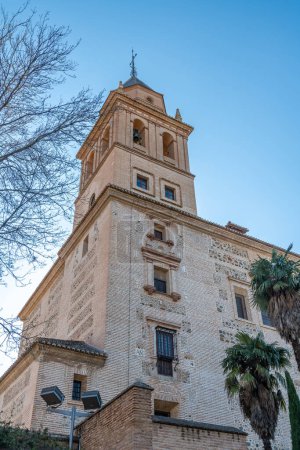 Foto de Granada, Granada - España - 30-01-2024: Junto a las antiguas murallas de la Alhambra, se levanta el majestuoso campanario de color arena de la iglesia de Santa María de la Encarnación junto a las palmeras y bajo un cielo azul - Imagen libre de derechos