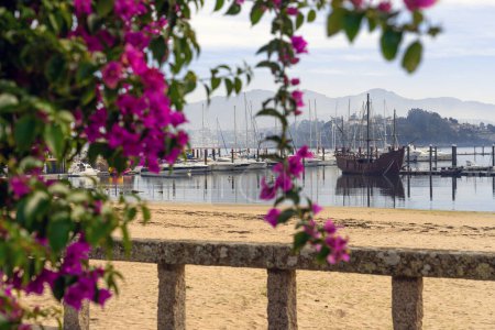 Téléchargez les photos : BAIONA, ESPAGNE - 23 OCTOBRE 2021 : caravelle du musée La Pinta dans le port de pêche de Baiona vue à travers une arche aux fleurs violettes. Pontevedra, Galice, Espagne. - en image libre de droit