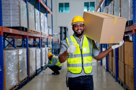 Photo for African American warehouse worker carry the box on his shoulder and show thumbs up to camera with smiling and his coworker check product on shelves in the background. - Royalty Free Image