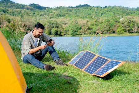 Téléchargez les photos : Homme asiatique assis sur le champ d'herbe et utiliser un téléphone mobile qui obtiennent de l'énergie à partir de cellules solaires pendant le voyage et le camping dans le parc national. - en image libre de droit