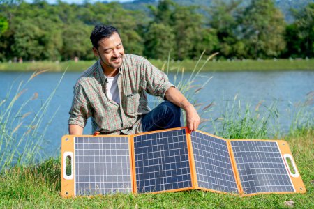 Asian man set up solar cells panel during camping in national park near the lake and he look happy.