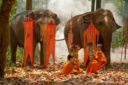 Photo for Two boys with Pee Ta Khon dress hold mask and talk together in front of elephant and traditional flags on background in forest. - Royalty Free Image