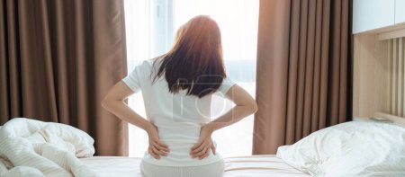 Foto de Mujer que tiene dolor de espalda durante sentarse en la cama en casa. hembra adulta con dolor muscular después de despertar debido al síndrome de Piriformis, dolor lumbar y compresión espinal. Concepto médico de salud - Imagen libre de derechos