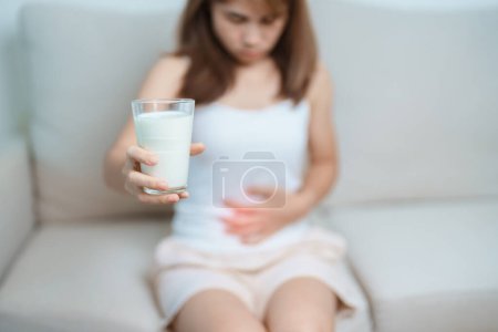 Téléchargez les photos : Intolérance au lactose et concept d'allergie au lait. femme tenir verre de lait et avoir des crampes abdominales et de la douleur quand boire du lait de vache. Symptômes maux d'estomac, intolérance aux produits laitiers, nausées, ballonnements, gaz et diarrhée - en image libre de droit