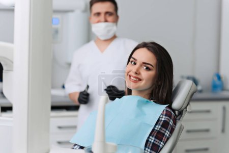 Photo for Satisfied dentist patient showing her perfect smile after treatment in a clinic box with dentist in the background - Royalty Free Image