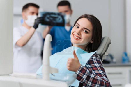 Porträt des Patienten, der in der Zahnarztpraxis den Daumen nach oben gibt, mit dem Zahnarzt und seiner Assistentin bei der Röntgenanalyse
