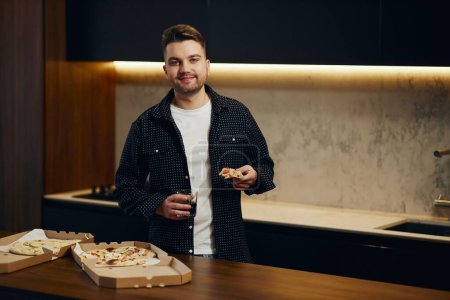 Téléchargez les photos : Handsome young man enjoying pizza in his kitchen. Unbelievably enjoys the taste. - en image libre de droit