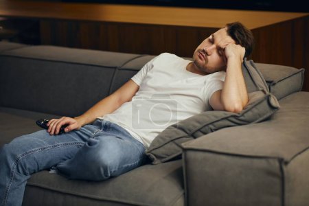 Téléchargez les photos : A young man in a white T-shirt, exhausted, fell asleep in front of the TV. Lazy man watching television at night alone. - en image libre de droit
