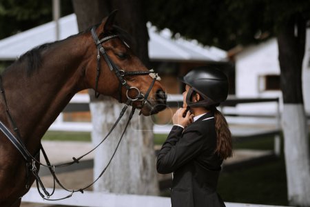 Foto de Attractive female equestrian in riding helmet looking at horse in horse club. - Imagen libre de derechos