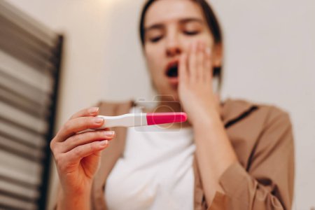 Foto de Young worried sad girl sitting in the bathroom and holding a positive pregnancy test in her hands. She is in shock, this is an unexpected pregnancy. The concept of different pregnancy emotions. - Imagen libre de derechos
