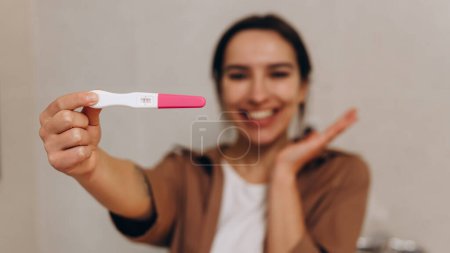 Foto de Happy young girl holding a positive pregnancy test in her hands. She is incredibly happy about this news. Happy motherhood and pregnancy concept. In the foreground is the test, focus on the test. - Imagen libre de derechos