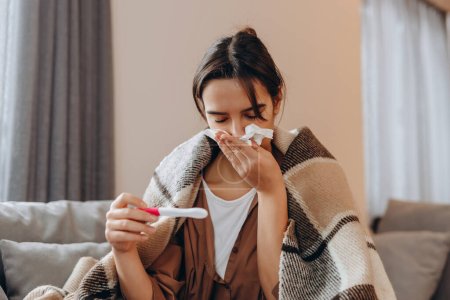 Coronavirus. Ill sick young woman blowing nose, coughing or sneezing in tissue, sitting on sofa covered with blanket at home, suffering from flu. Cold and fever concept