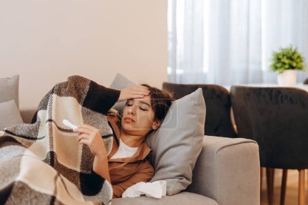 Una mujer enferma mide su temperatura mientras está sentada en el sofá y se cubre con una manta.