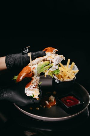 Foto de Desgarrado en la mitad hamburguesa de pescado en las manos con patatas fritas y la pausa en el plato negro - Imagen libre de derechos