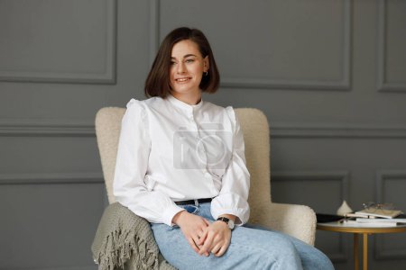 Téléchargez les photos : Portrait d'une femme d'affaires souriante portant une chemise blanche et un jean assis les mains croisées sur les genoux dans une chaise beige avec un plaid gris sur un fond mural gris - en image libre de droit