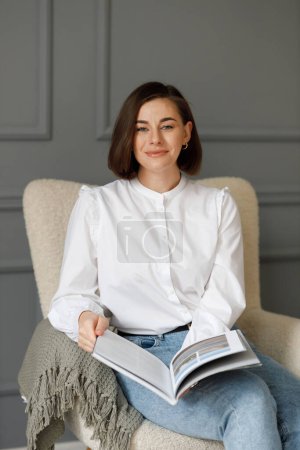 Téléchargez les photos : Jeune belle femme d'affaires souriante portant une chemise blanche et un jean avec un magazine à la main, assise sur une chaise beige avec un plaid gris devant un mur gris dans la chambre - en image libre de droit