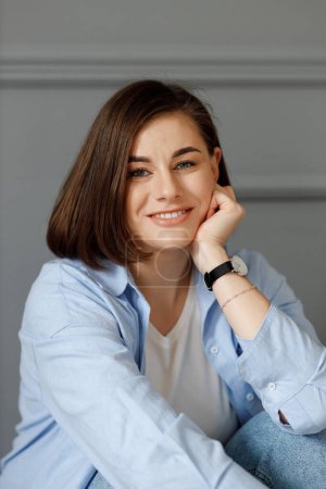 Photo for Portrait of a young beautiful girl which is holding a hand with a watch and a bracelet on her chin, wearing a white t-shirt and a blue shirt on a gray background - Royalty Free Image