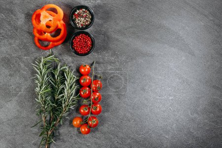 Una disposición estética de ramitas de romero fragante, tres anillos de pimiento rojo, una rama de tomates cherry y dos tazas negras con guisantes de pimiento mixtos y rojo en una mesa de cocina de piedra