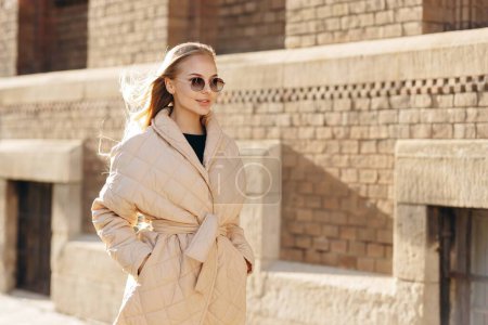 Téléchargez les photos : Une jeune femme moderne avec des lunettes de soleil sur les yeux porte un pull noir et un imperméable beige et se tient sur le fond d'un beau bâtiment - en image libre de droit